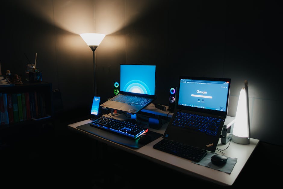 A sleek gaming desk with two laptops, RGB keyboard, and ambient lighting for a modern tech setup.
